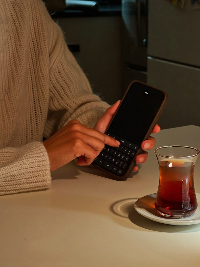 Hände einer Frau im Strickpullover, die abends am Küchentisch auf dem Handy eine Nachricht tippt, daneben ein Glas türkischer Tee